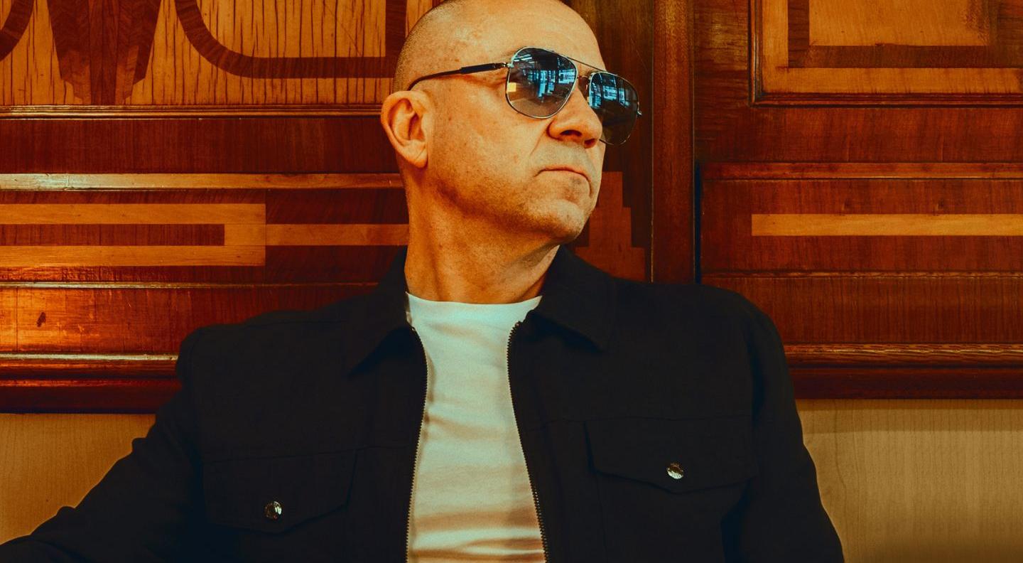 Kenny Thomas wears a black jacket and round sunglasses. He is bald, and leans back against wood panelling, looking off to his left