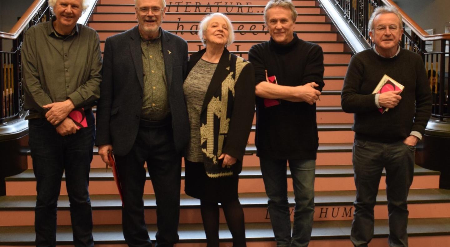 The Lost Poets standing in a line on the staircase of the National Library of Scotland with the word Literature visible on the stairs behind their heads. Other words are only partly visible behind their bodies. They are shown in full length smiling and looking at the camera and are all holding books with a pink cover. 