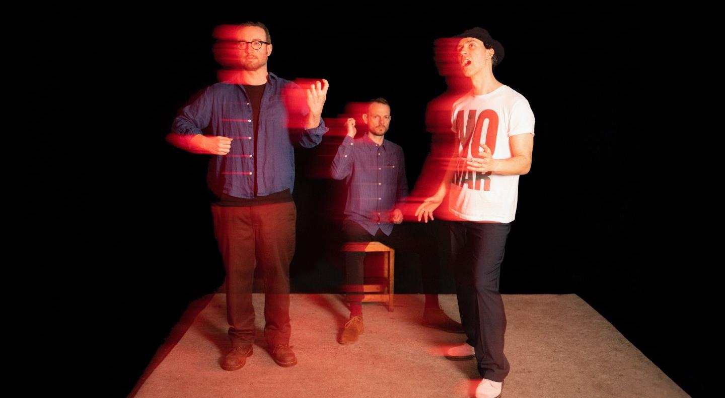 Three men stand on a darkened stage lit in red, the image is blurred as though in motion