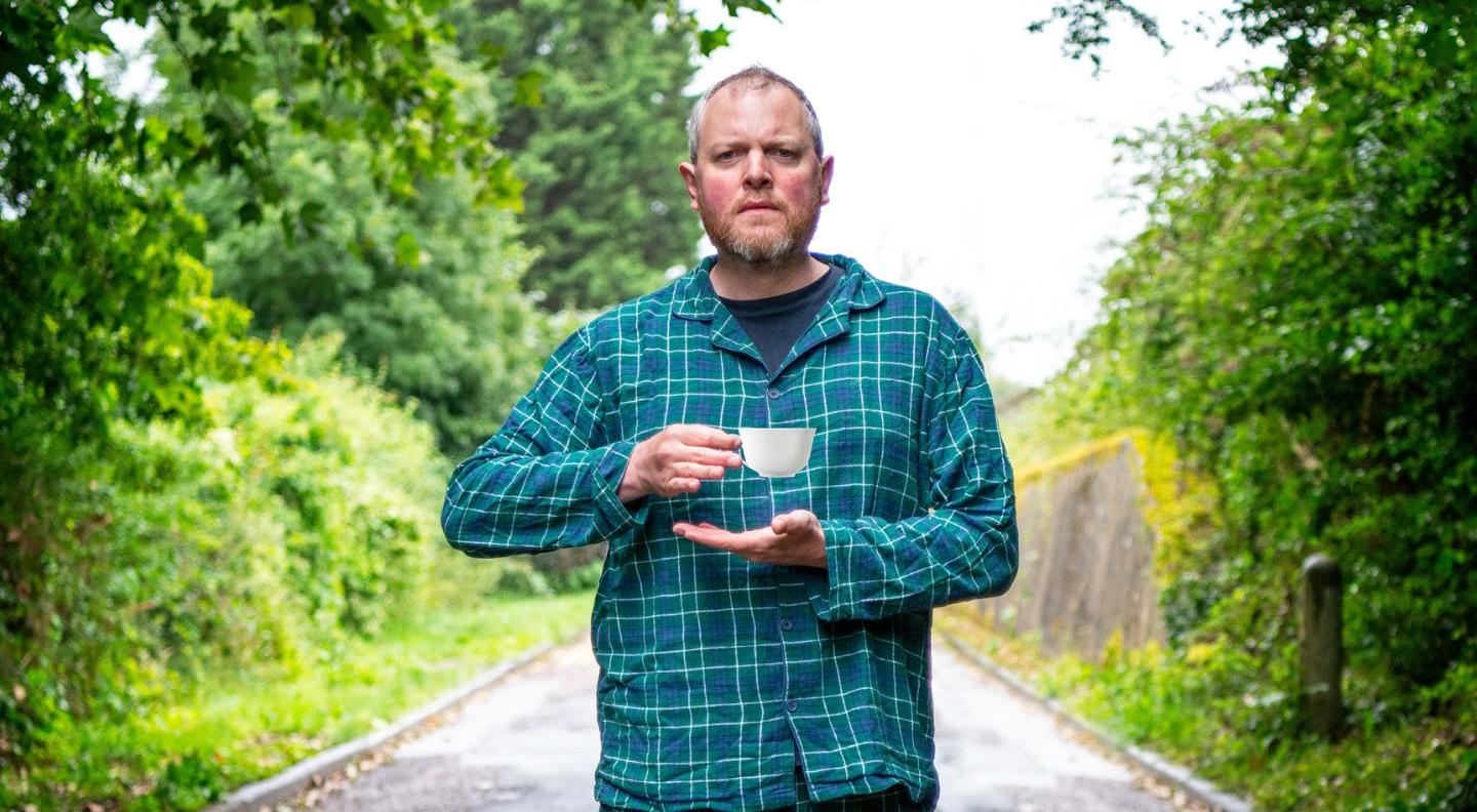 Miles Jupp, a white bald man, stands in a road lined by trees. He's wearing blue tartan pyjamas and holding a cup of tea
