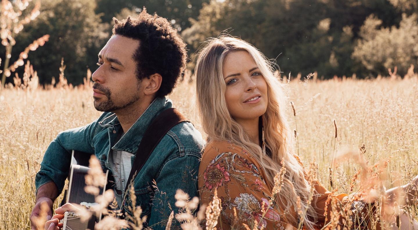 A mixed race man and white woman with long blonde hair sit back to back in a sunny field of corn