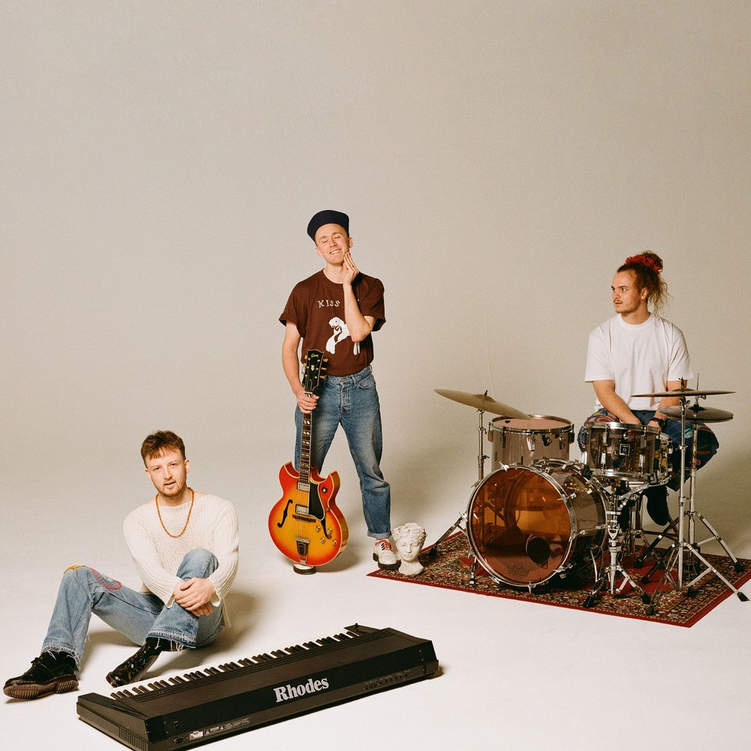 Three young white men dressed casually sit or stand behind their instruments - a keyboard, guitar and drum kit