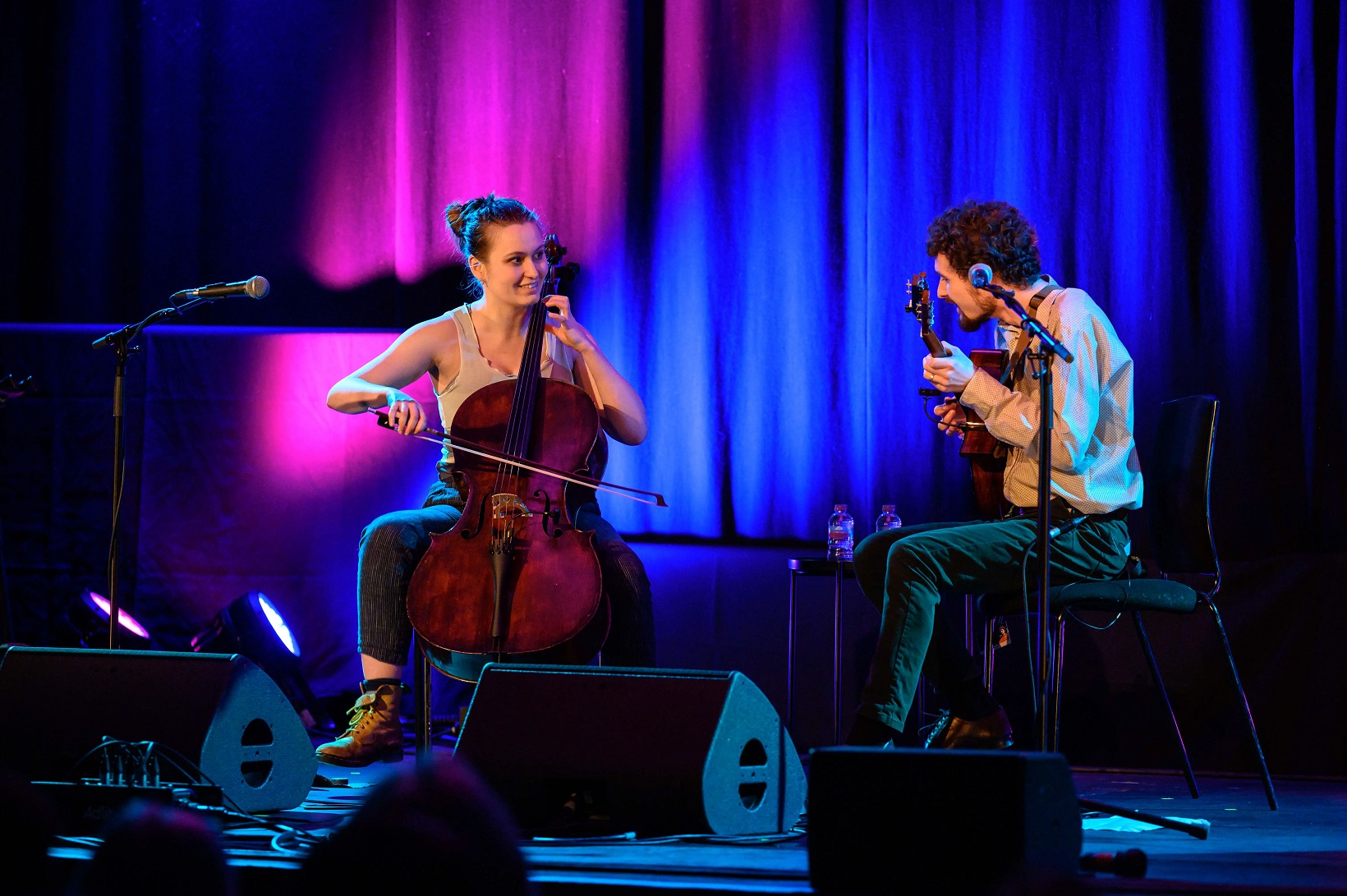 A young white woman and white man play the cello and guitar facing each other on a stage lit with pink and blue lights