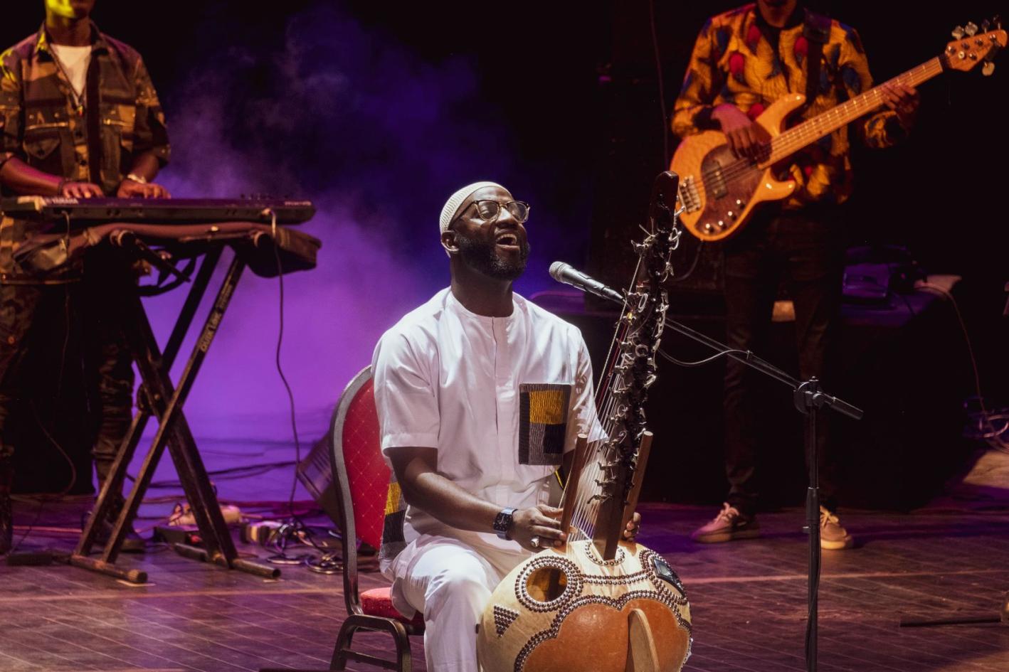 A black man sits on the edge of stage playing the kora, a string instrument. A keyboard player and guitarist can be seen in the background