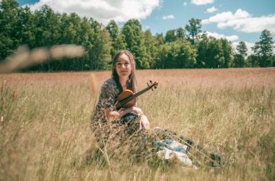 A young white woman in a floaty summer dress sits in a field of yellowing grass holding a violin