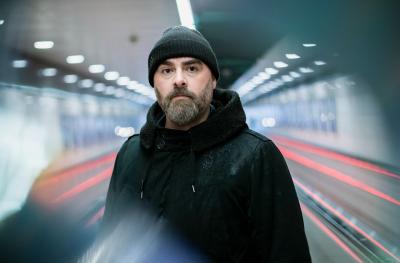 A white man wearing a dark beanie and coat stands in front of a background which gives the impression of fast-moving traffic