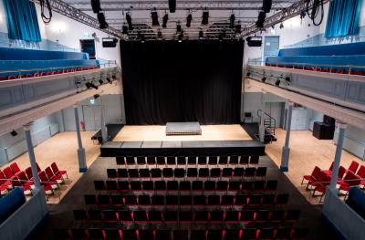 A view from above of a stage with a black backdrop ,rows of red seats in front, seats on a gallery and a lighting rig