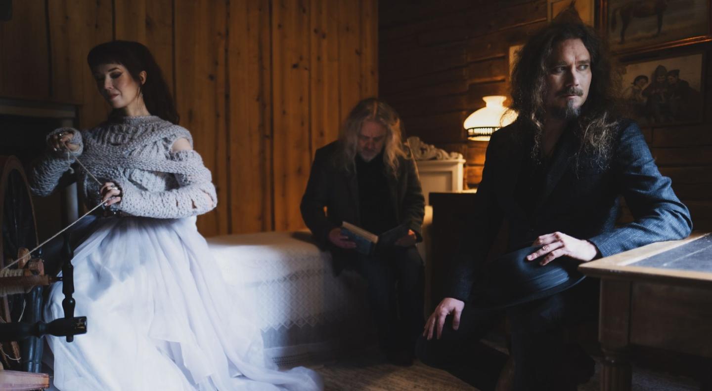 A woman in a white wedding dress spinning yarn while look off left behind her sit two menu with long hair and beards looking wistful