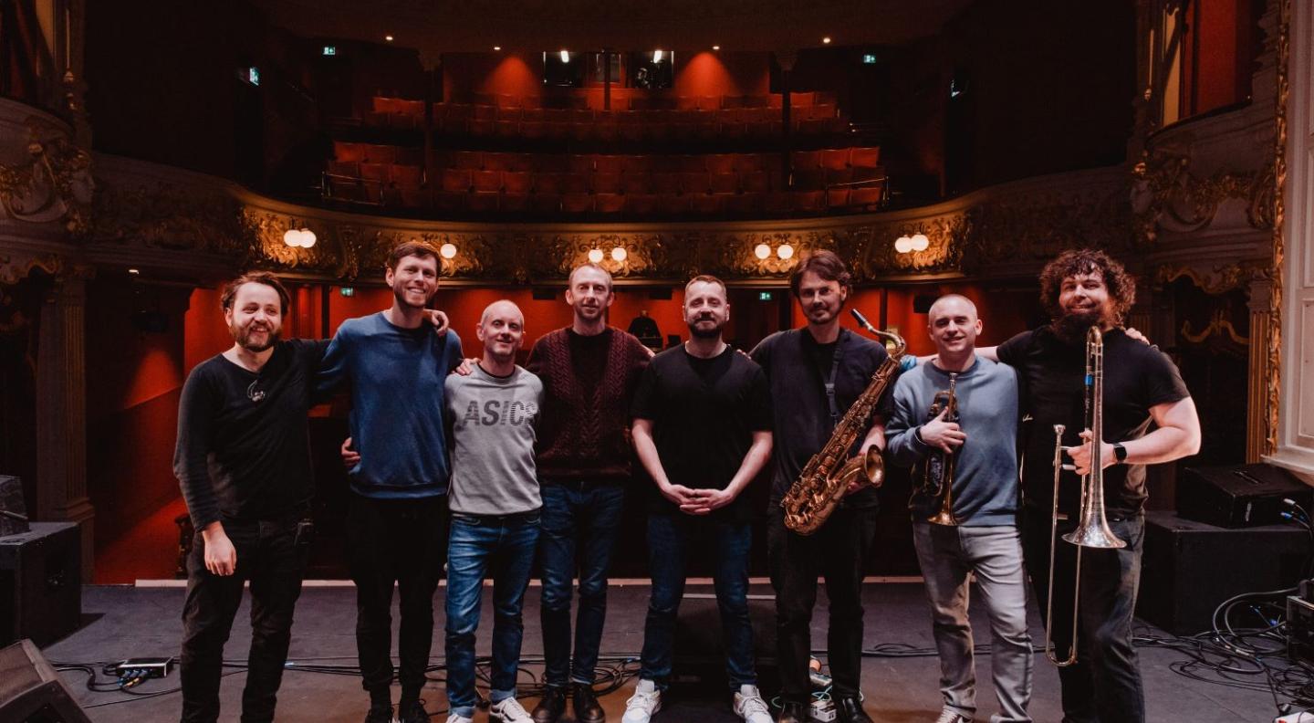 Eight white men stand on stage in a line smiling with a backdrop of an empty, ornate auditorium. Three of them hold brass instruments.