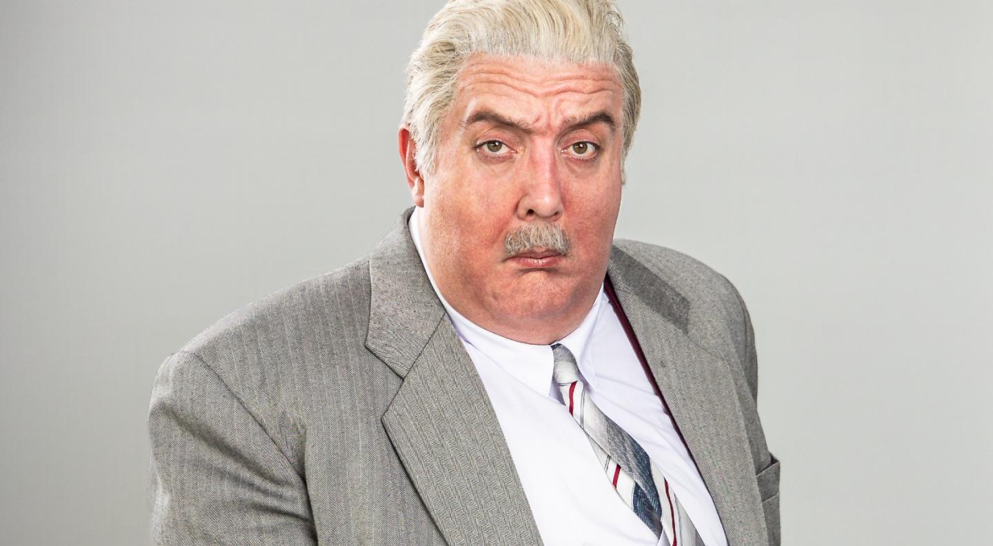 An overweight older man with white hair with a sour look on his face. He wears a grey suit, patterned tie and white shirt and is staring out at the viewer.
