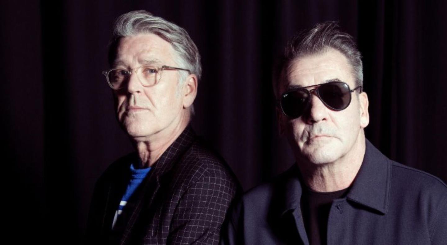 A midshot of two middle-aged white men, one wearing sunglasses, looking at the camera with serious expressions