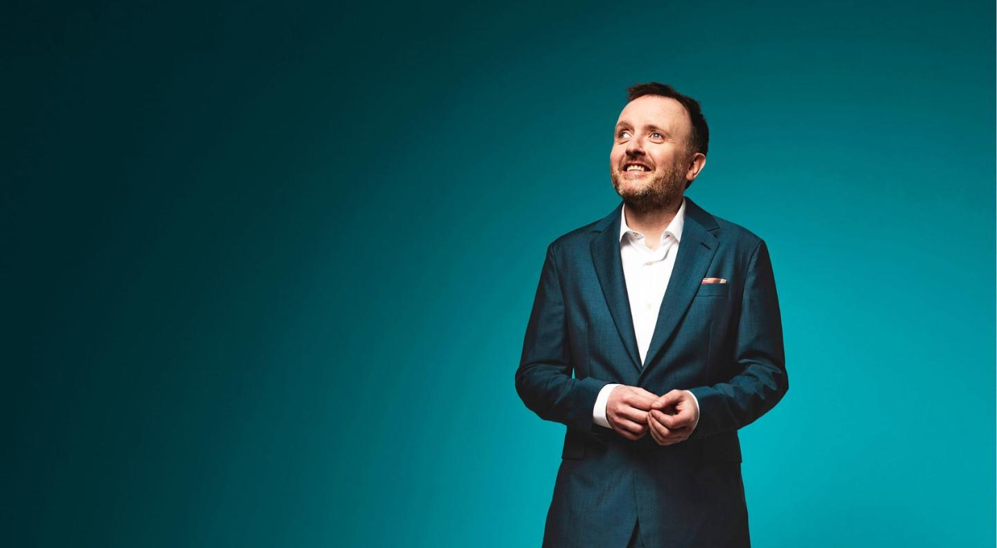 an image of comedian Chris McCausland against a green background, wearing a dark grey suit