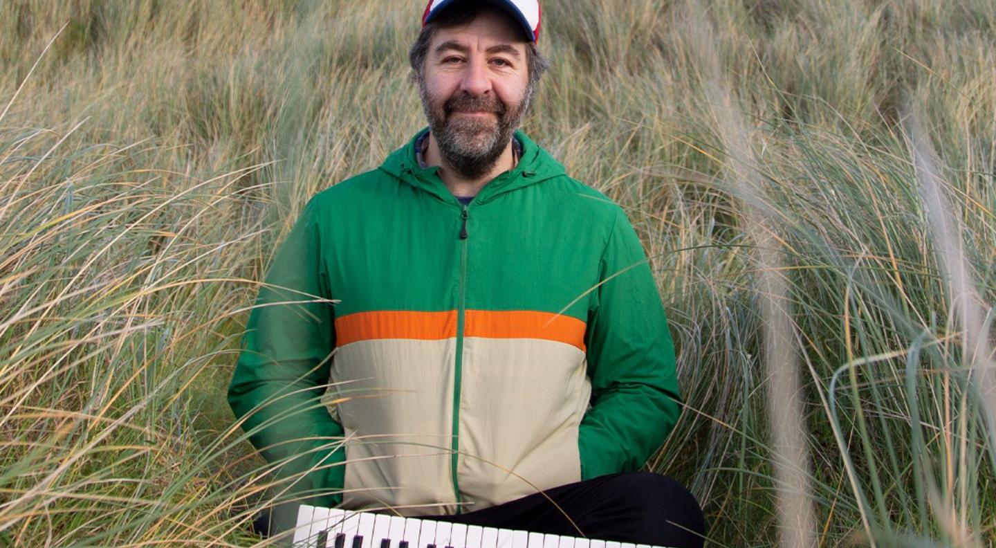 a man stands in a field of grass wearing a green jacket and white baseball cap, with a keyboard in front of him