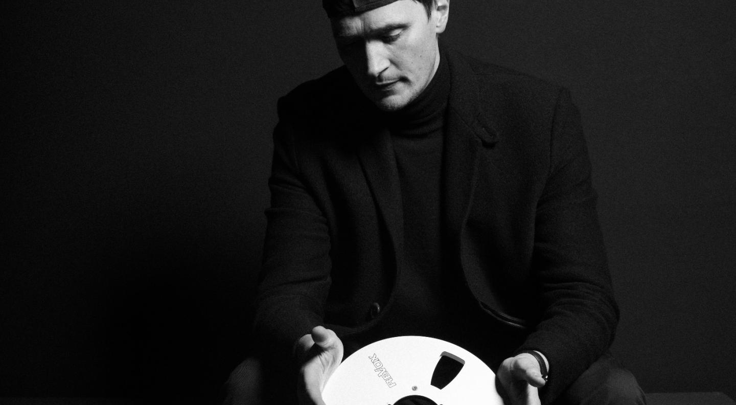 A black and white image of Erland Cooper - a white man wearing dark clothing and a flat cap backwards - looking down at a silver film reel he holds in his hands. The photo is credited to Andrea Fumana