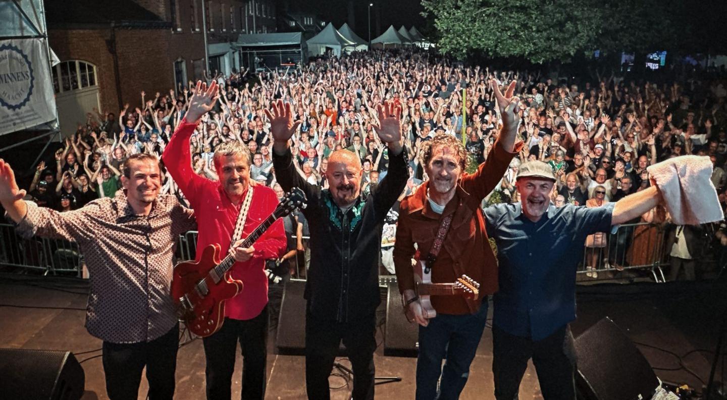 An image of the band on stage, with the audience at their backs, all looking into the camera