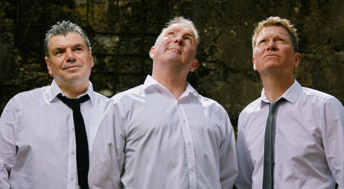 The three members of Frost stand looking up at the sky. They all wear white shirts, two wear black ties