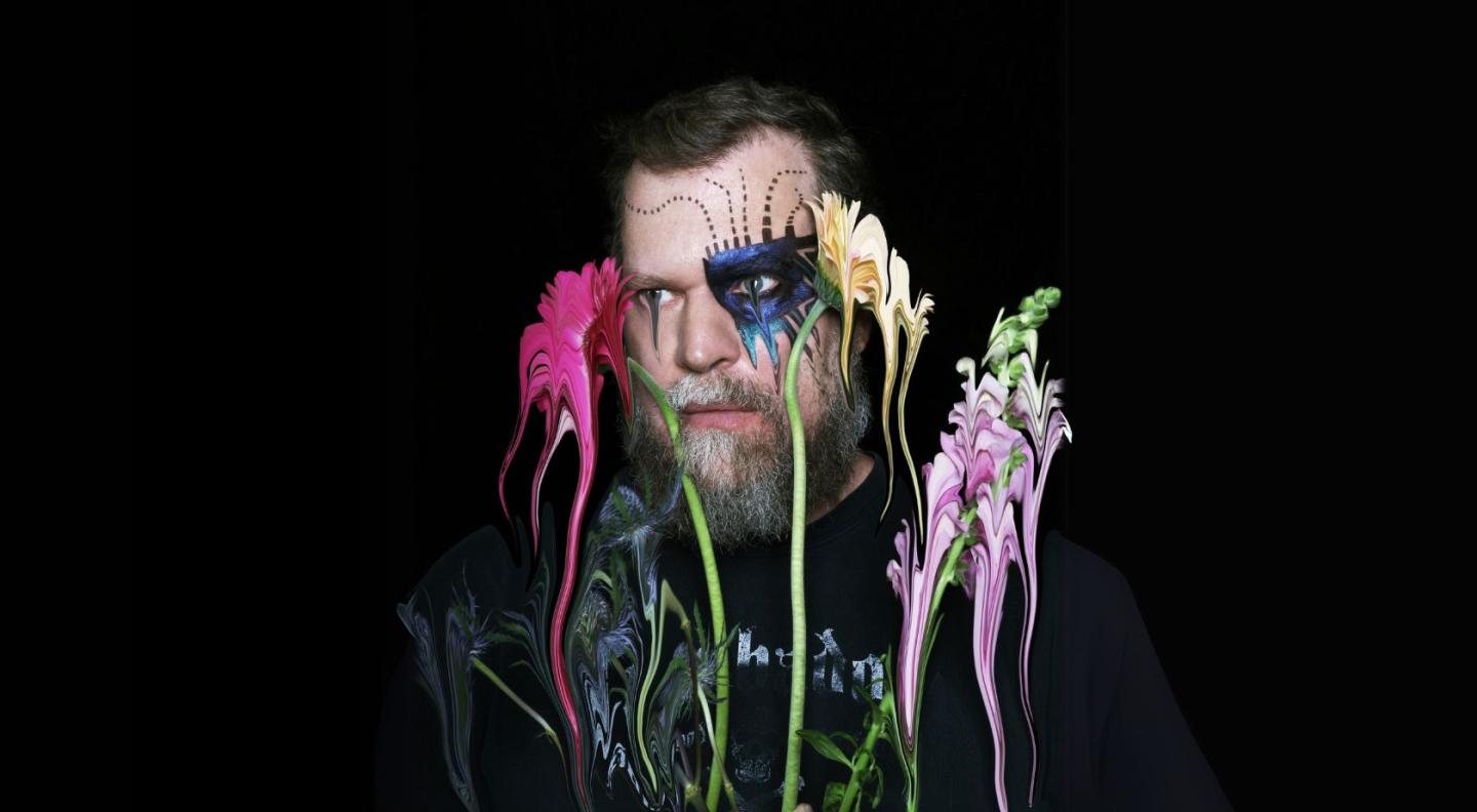 John Grant in front of a black background with artificial flowers and colours superimposed over his face.