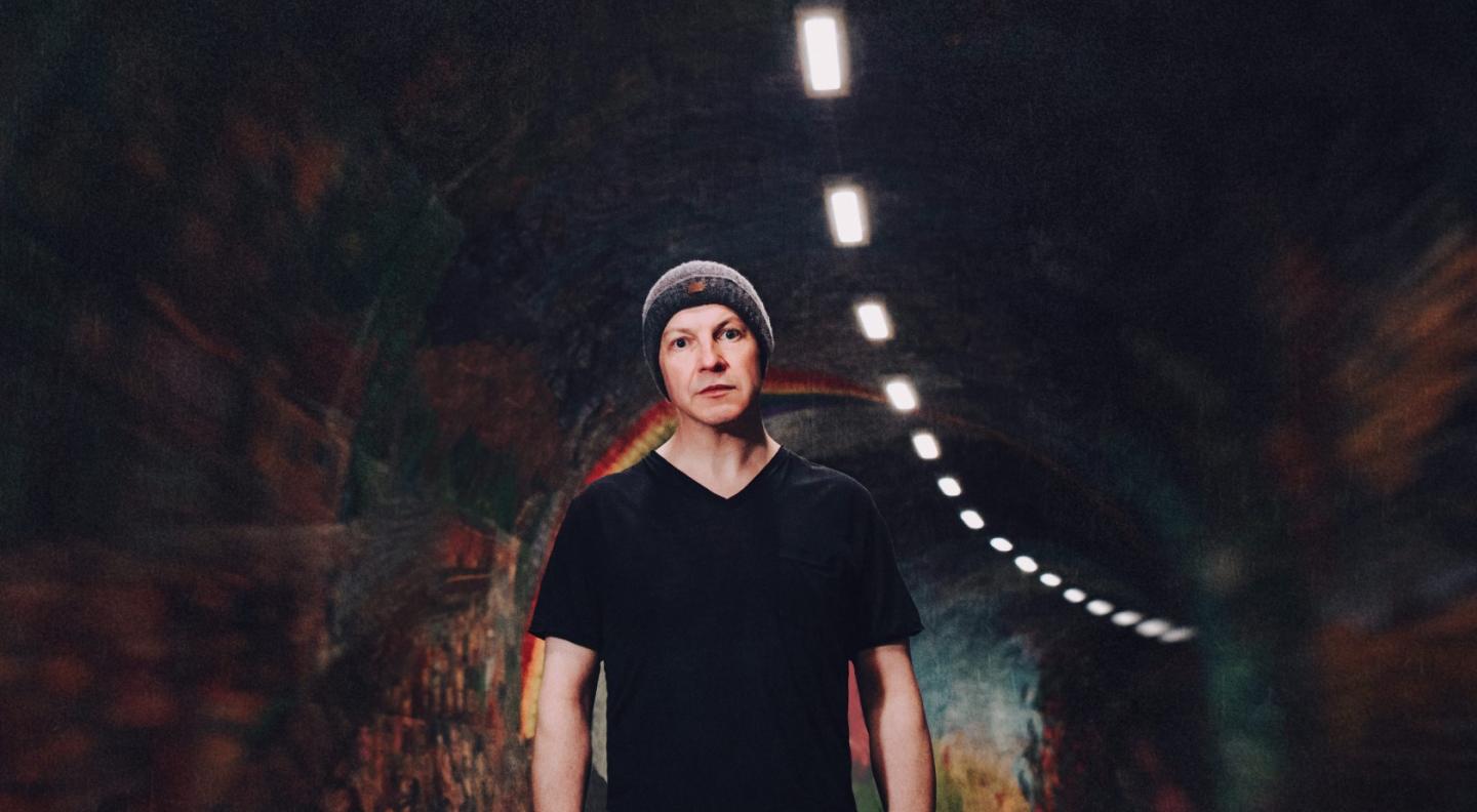 John McCusker - a white man wearing a dark t-shirt and beanie hat - stands in a tunnel with curved walls looking out at the viewer. A row of lights in the ceiling runs overhead into the distance