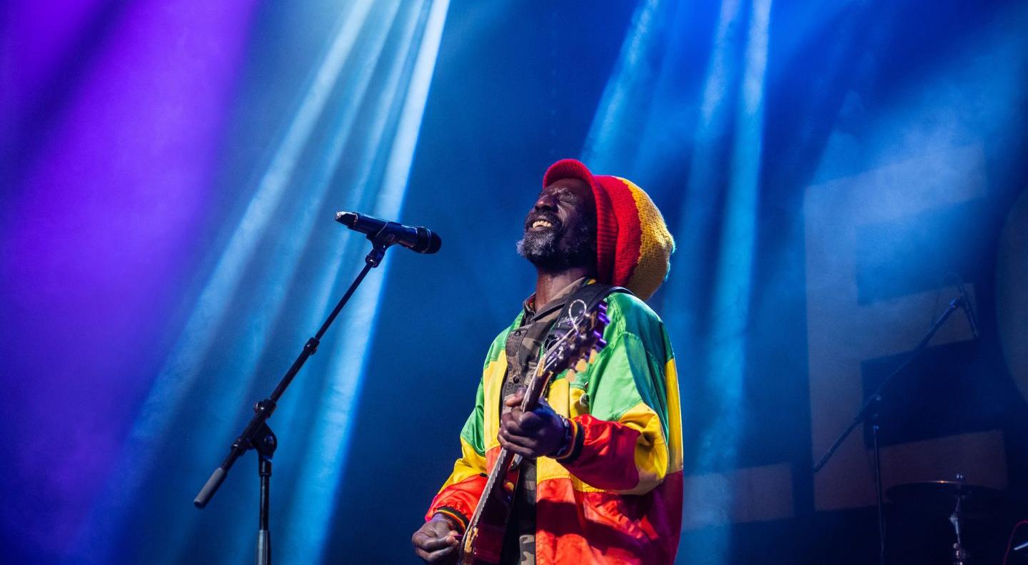 an image of the performer on stage, wearing a large knitted hat and a brightly coloured jacket