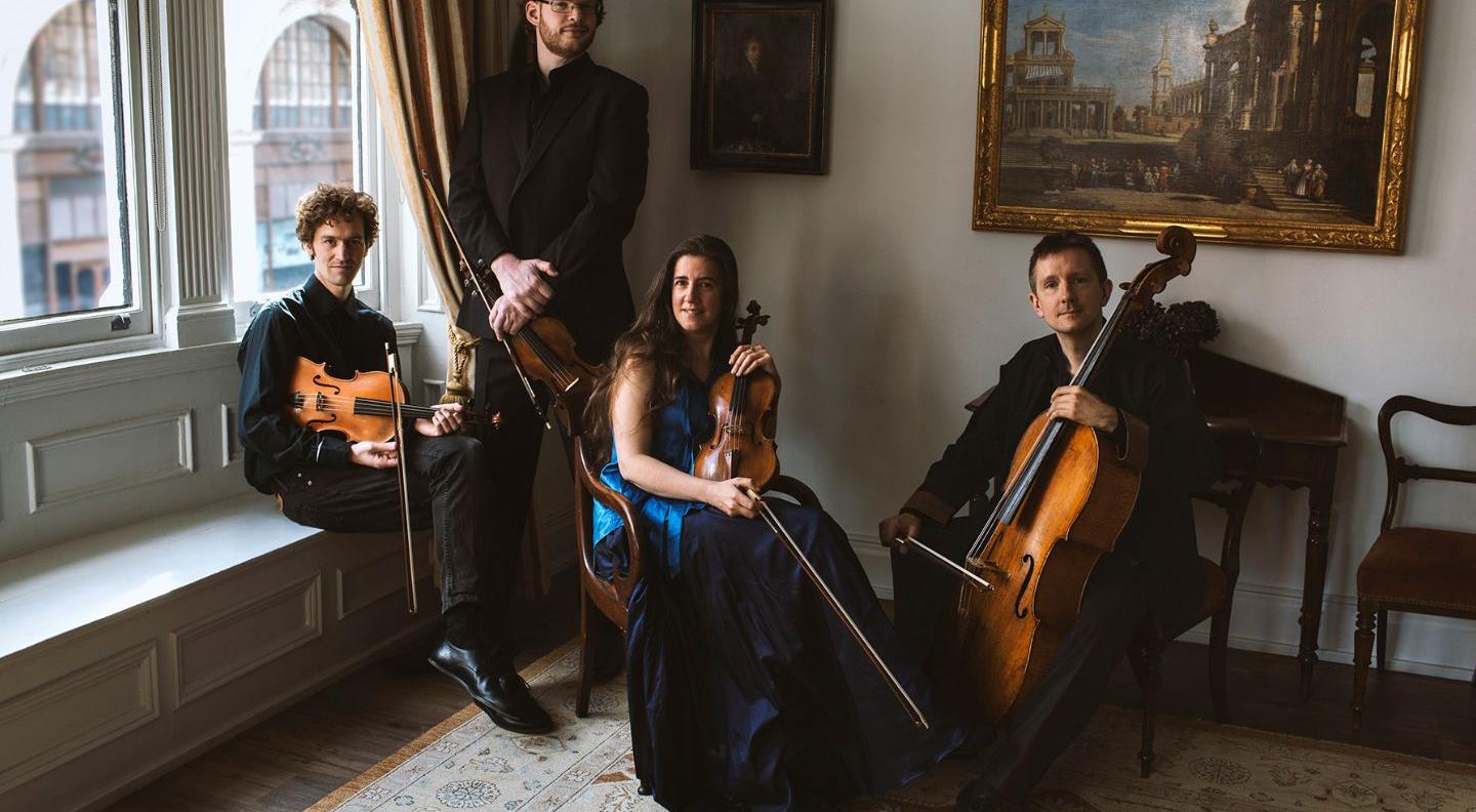 Four musicians sit in an ornately decorated Georgian room, holding their instruments. A man and a woman hold violins, one man has a viola, and one man has a cello 
