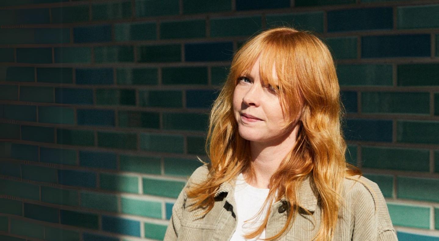 Lucy Rose, a female singer with ginger hair, standing in front of a green brick wall