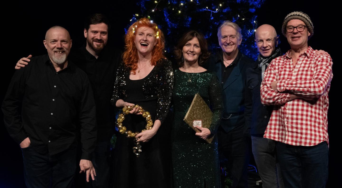 Phil Cunningham stands alongside 6 Christmas Songbook colleagues in front of a Christmas tree. 