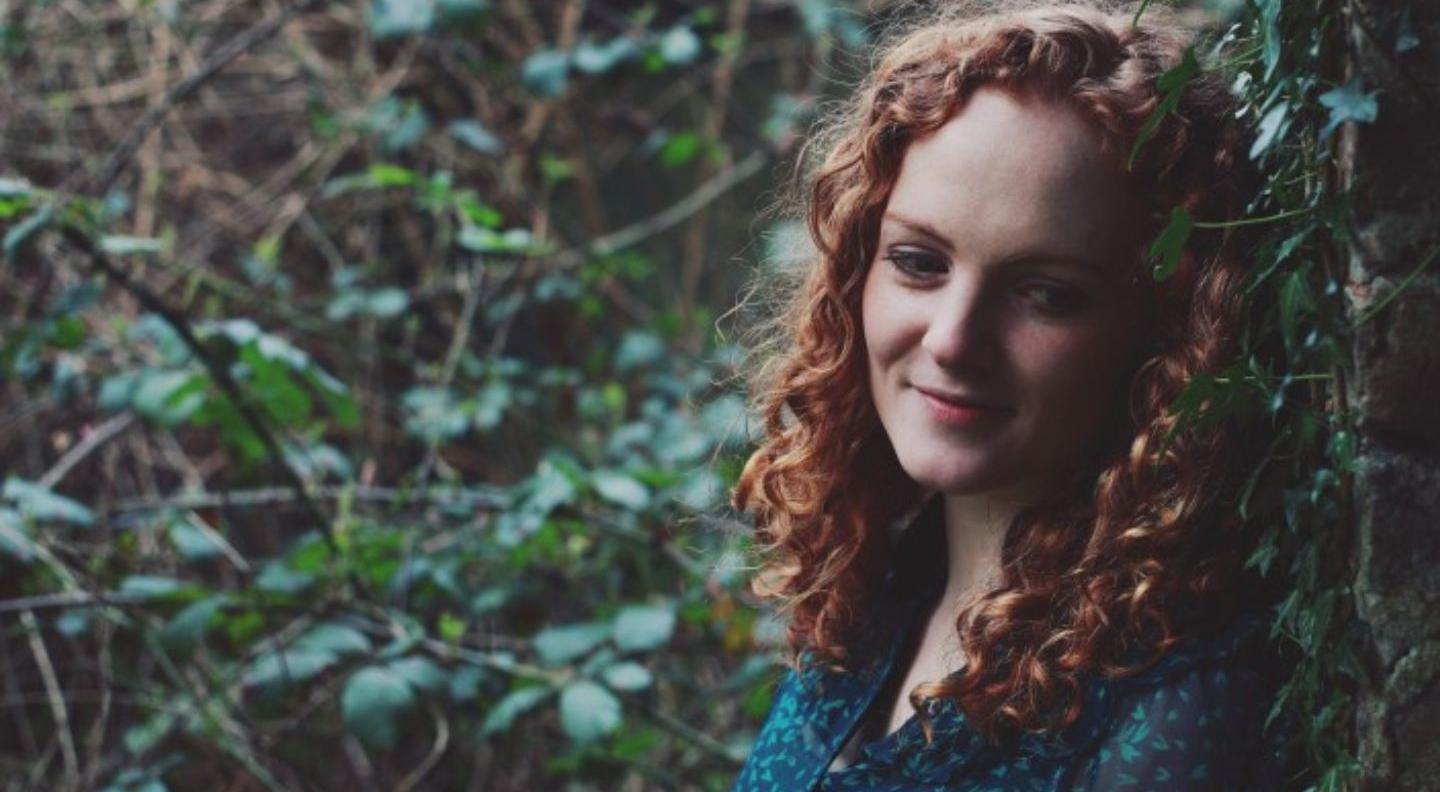 An image of Robyn, a white woman with curly red hair, leaning against a tree in the middle of a forest