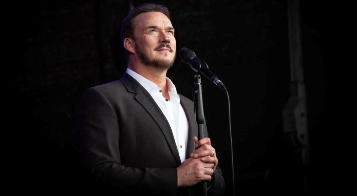 A white man with dark hair and facial hair wears a dark jacket and white shirt. He stands, looking upwards, holding a microphone stand