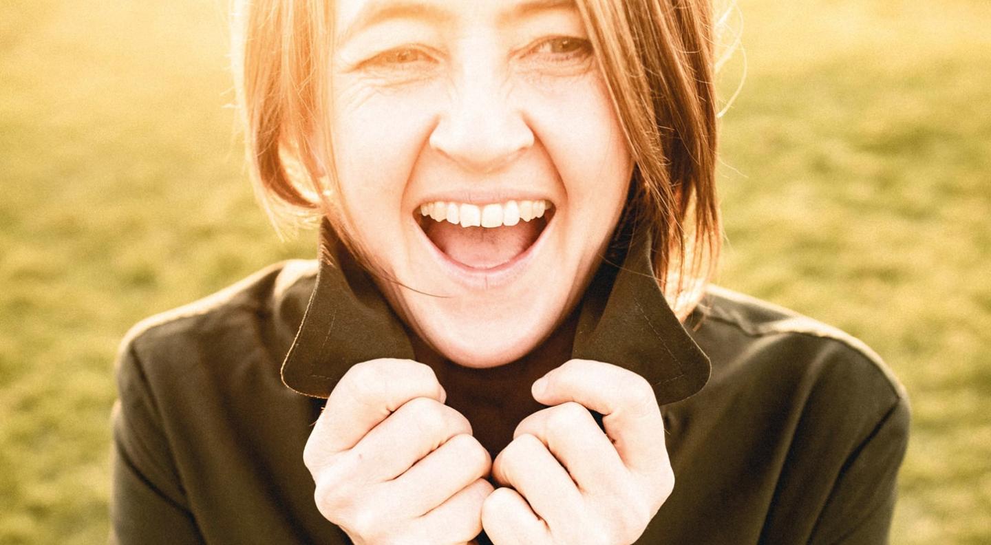 A full colour mid shot of Karine Polwart laughing with grass behind her and sunshine around her face