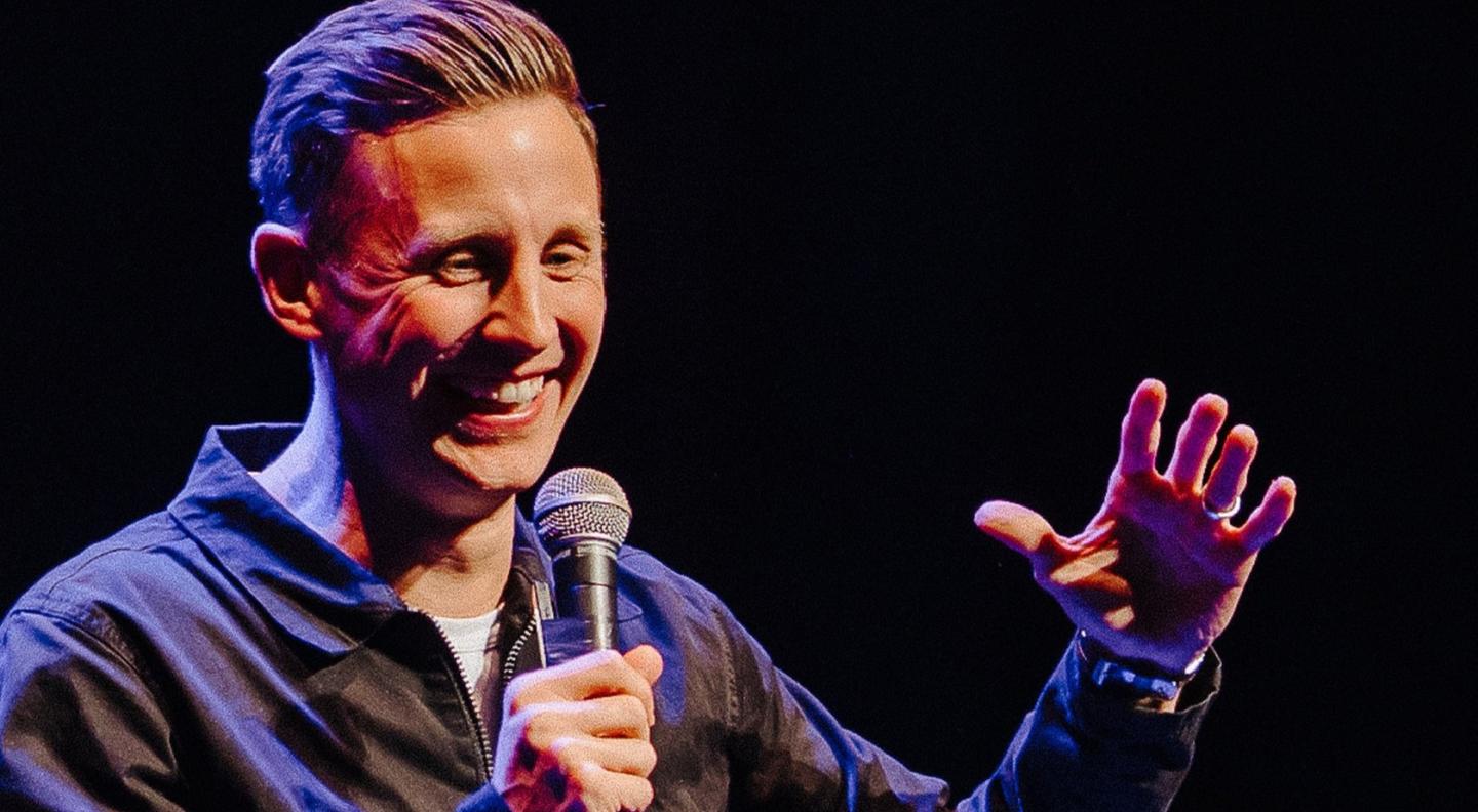 A white man with sandy brown hair is laughing into a microphone