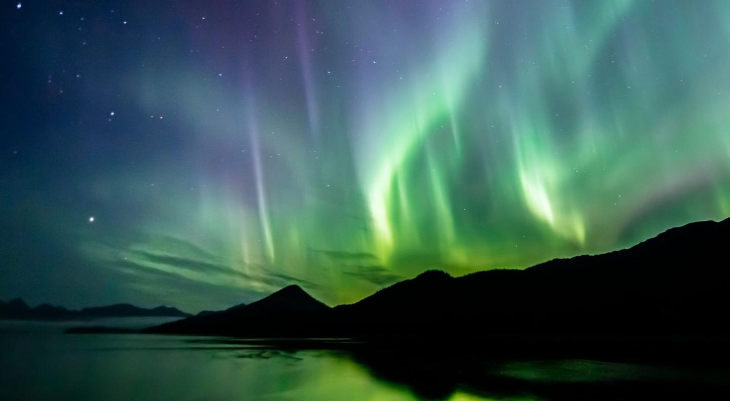 an image of northern lights over a dark landscape. The bright blues, greens, and purples are reflected in the water below