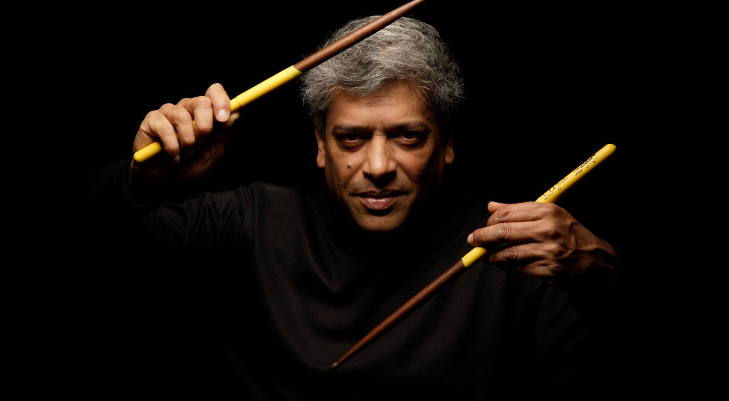 an image of Trilok Gurtu, a man with grey hair, holding two drumsticks against a black background