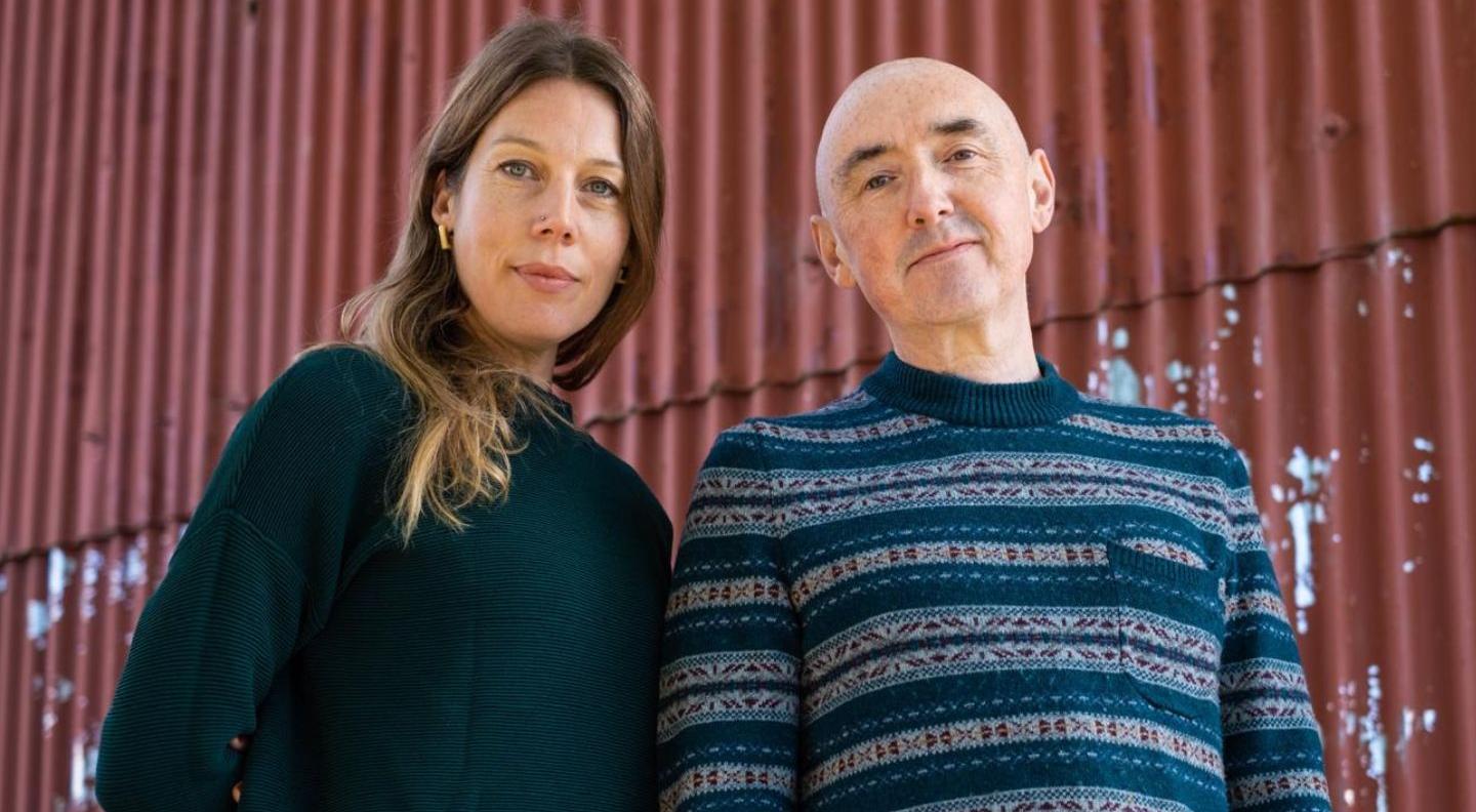 an image of the band snowgoose, a man and a woman, standing in front of a corrugated iron wall. They are both white, the woman has long brown hair and the man is bald.