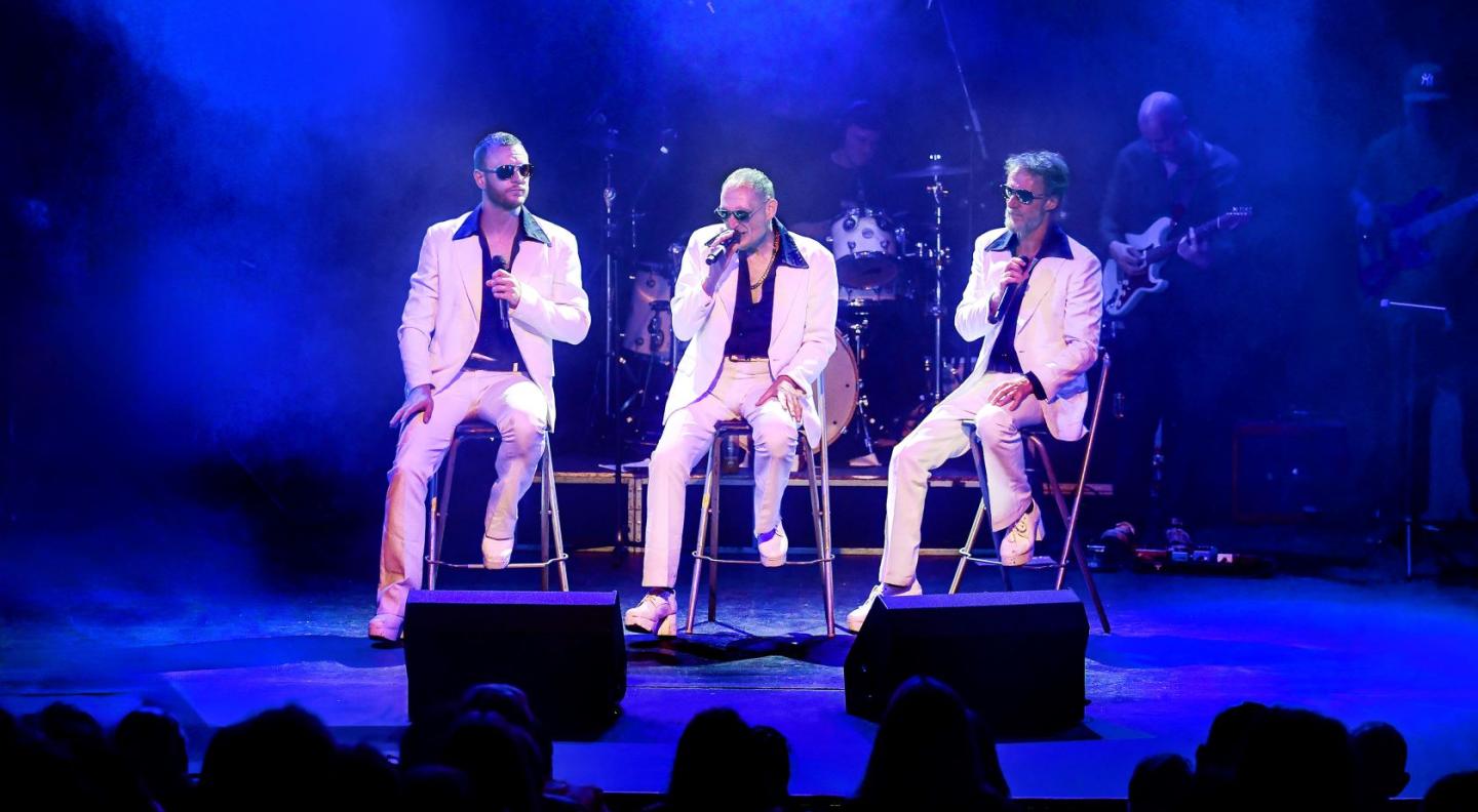 Three white men in white suits sit singing on bar stools on a blue lit stage