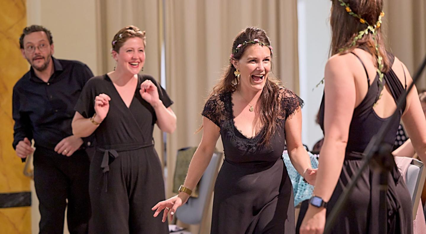 Three white women with flower wrathes in their hair laugh and look happy as they perform opera. A mischievous looking white man stands behind them